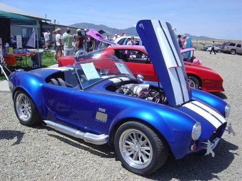 1986 Ford Bronco II - A blue AC Cobra replica is displayed with its hood open at a car show, with other vehicles and people in the background.