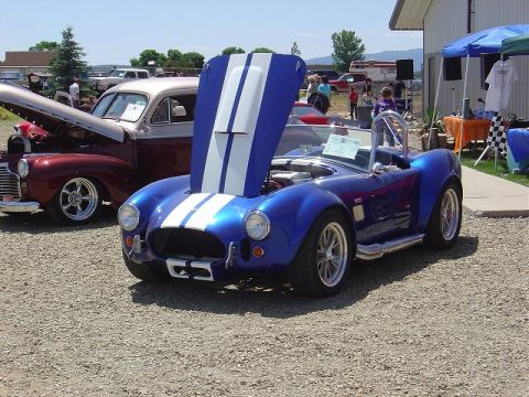 1986 Ford Bronco II - A blue AC Cobra replica is displayed with its hood open at an outdoor event, alongside another vintage vehicle.