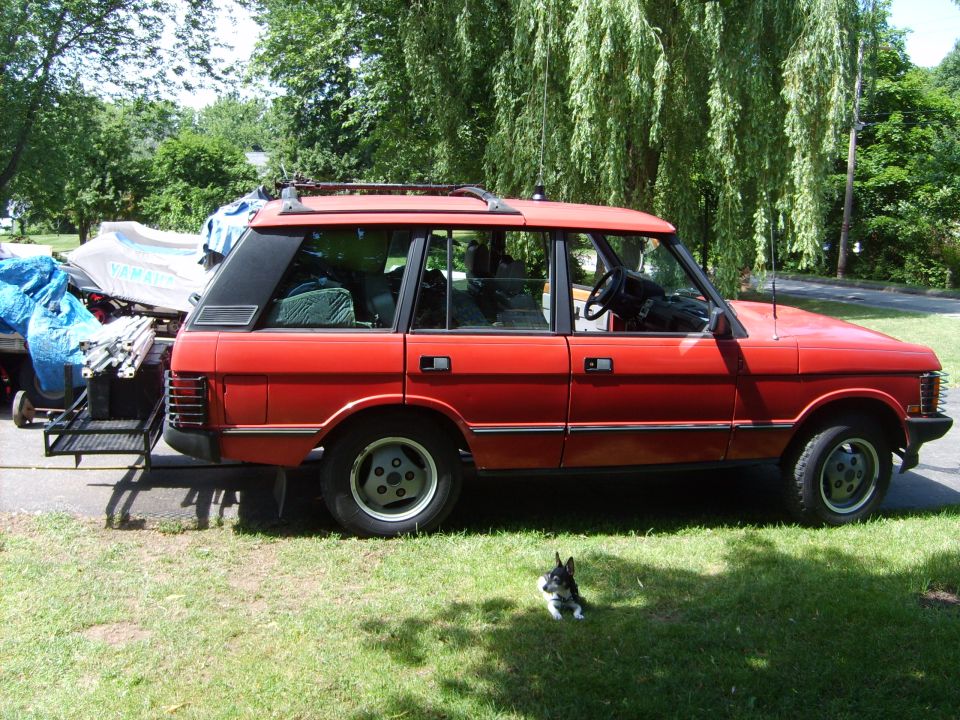 Red 4-door SUV with roof rack parked on grass beside a covered watercraft trailer; small dog lying in front