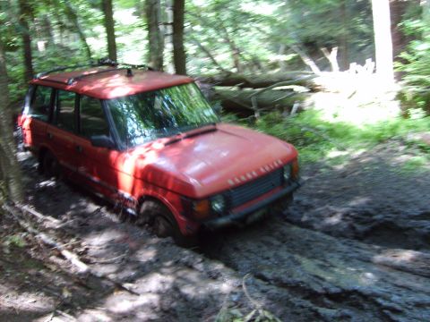 1995 Ford Bronco - A red Range Rover is stuck in deep mud on a trail in a forest.