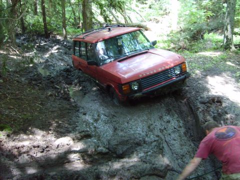 1995 Ford Bronco - A red Range Rover is stuck deep in mud on a wooded trail, with a person nearby attempting to assist.