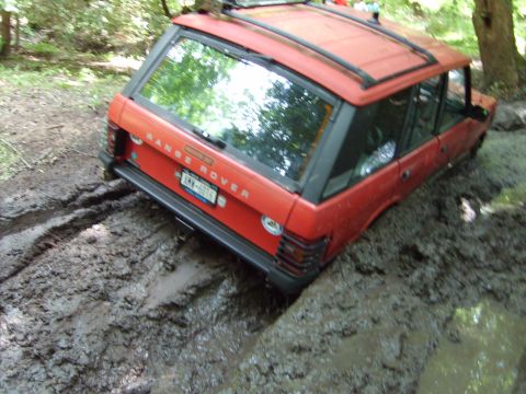 1995 Ford Bronco - A red Range Rover is stuck in thick mud on a wooded trail, its tires sunk deep into the mire.