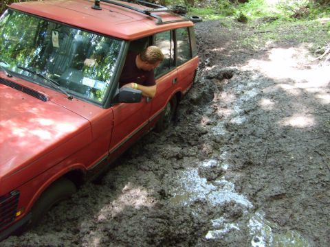 1995 Ford Bronco - A red Range Rover is deeply stuck in thick mud on a wooded trail, with the driver looking out the window.