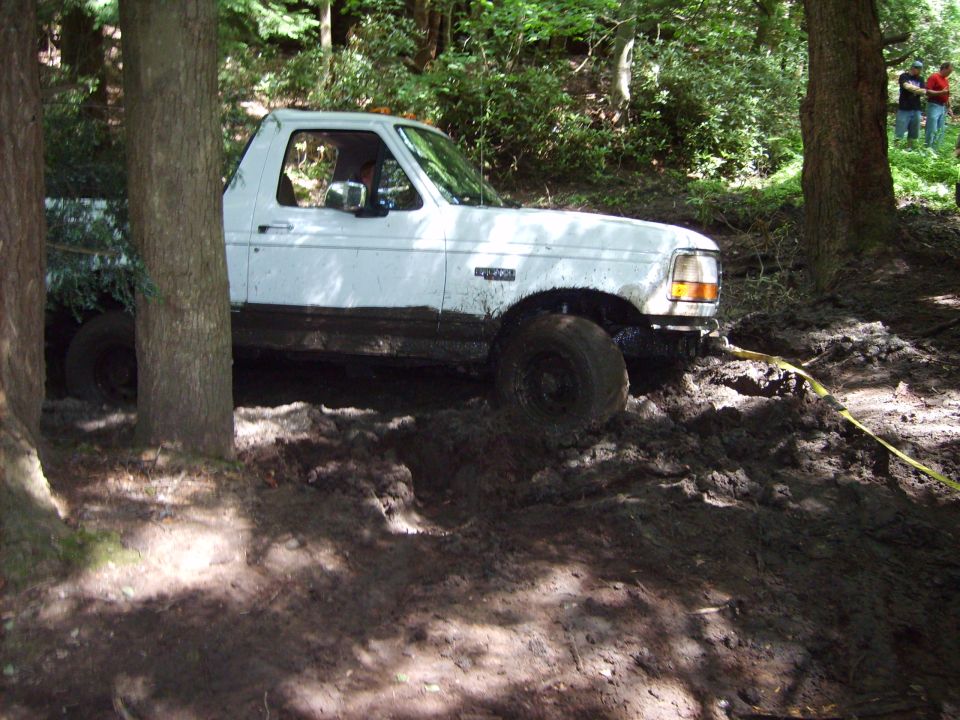 White Ford truck stuck in mud, side view, with trees and people in background