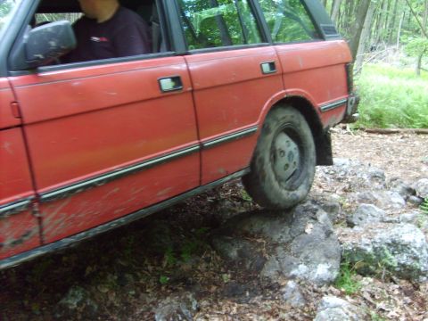 1995 Ford Bronco - A red SUV navigates a rocky path in a wooded area, with its tires on large stones.