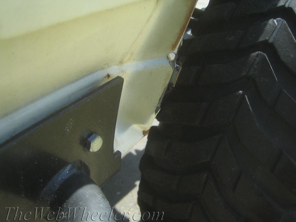 Close-up of a cream-colored vehicle body panel with rust spots and a black metal bracket bolted to it, next to a large, knobby tire