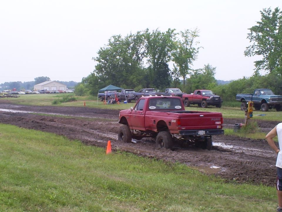 Red pickup truck with lifted suspension driving through mud. Other trucks and tents are visible in the background