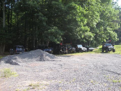 1991 Ford Bronco - A collection of off-road vehicles are parked in a gravel lot, with a dense forest in the background.