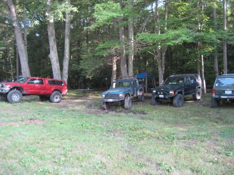 1991 Ford Bronco - Several off-road vehicles are parked in a clearing surrounded by trees, with a picnic table visible in the background.