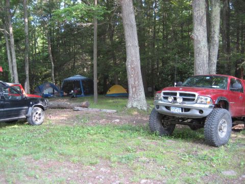1991 Ford Bronco - A red pickup truck with oversized tires is parked at a campsite with tents in the background.