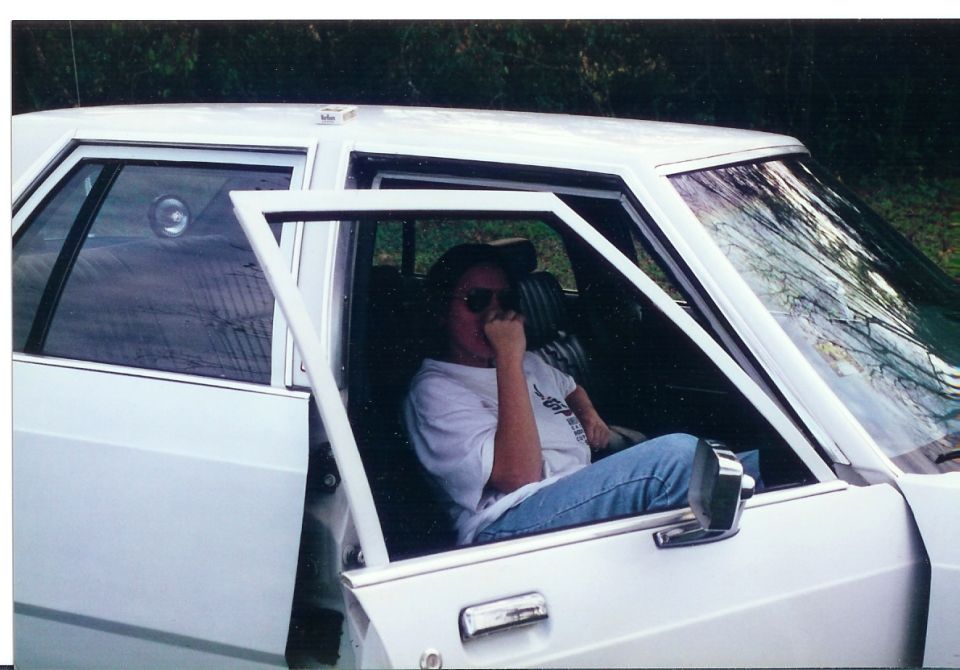 Person sitting in a white 1980s sedan with the driver's door open