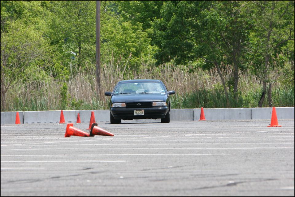Black sedan driving on a paved surface with orange cones. License plate reads PBJ-264