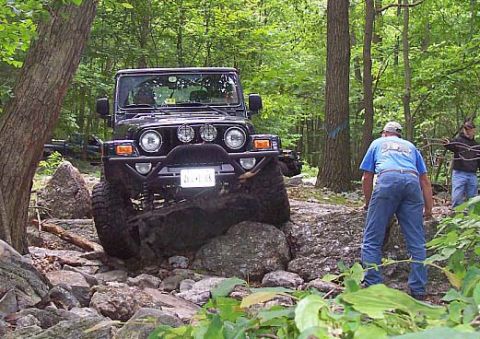 2003 Jeep Wrangler - A black 2003 Jeep Wrangler tackles a challenging rocky path in a wooded area, with two people observing.