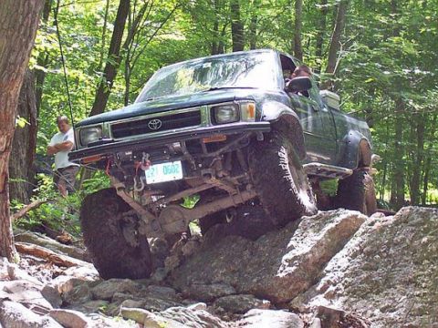 2003 Jeep Wrangler - Modified Toyota pickup truck tackling a difficult off-road obstacle course in a wooded area.