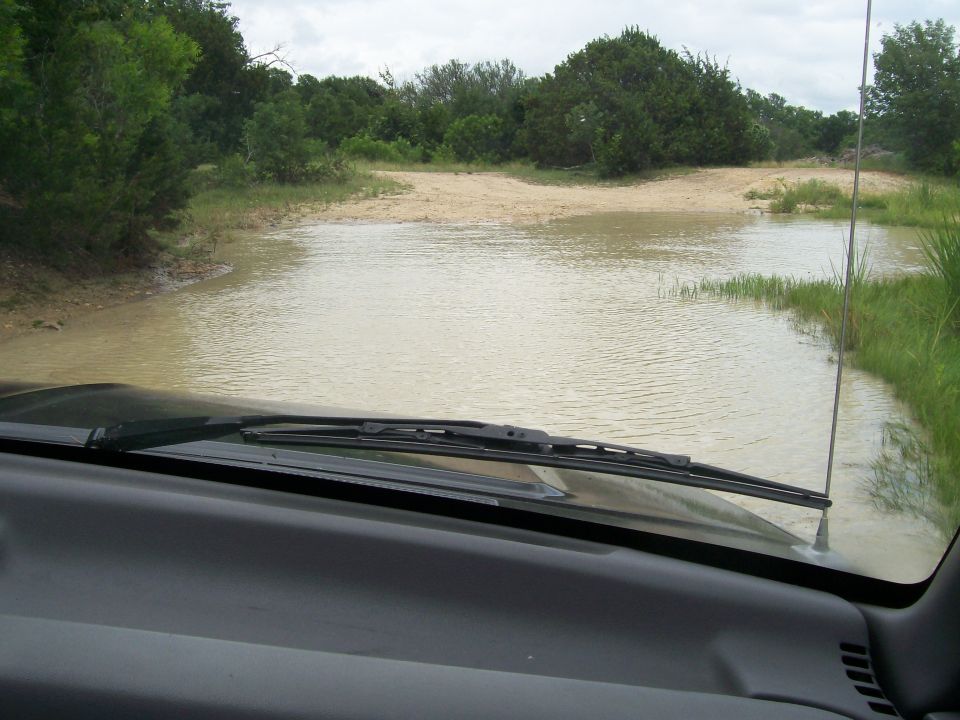 POV from vehicle interior facing a flooded sandy water crossing with brush and trees.
