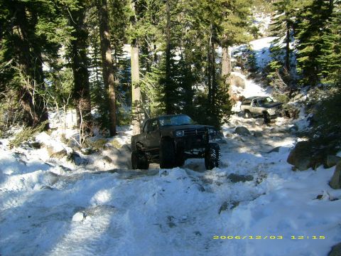 1986 Ford Bronco - Modified trucks navigate a challenging snowy and rocky path in a wooded area.