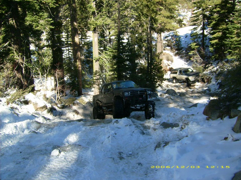Two modified trucks driving on a snowy, rocky trail through a forest