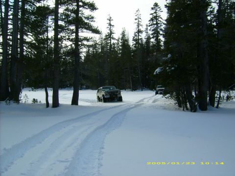1986 Ford Bronco - Two SUVs navigate a snow-covered forest road, leaving tire tracks behind.