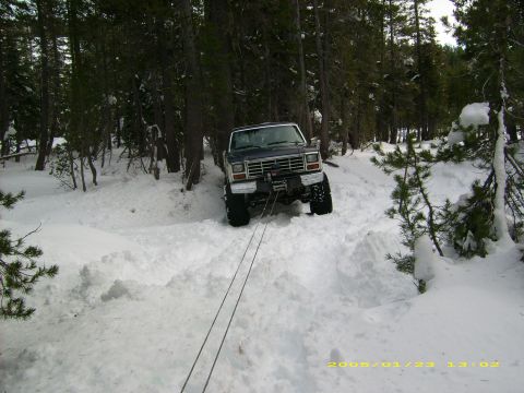 1986 Ford Bronco - A dark blue 1980s Ford Bronco is winching itself through a snowy forest trail.