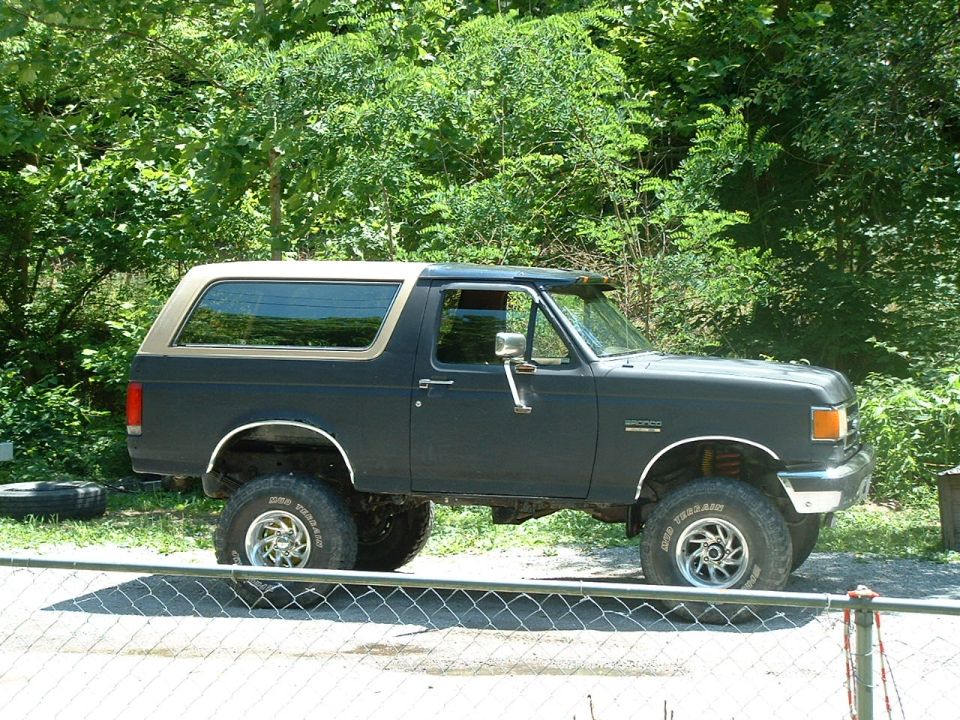 Dark gray 1991 Ford Bronco with a tan top, lifted on off-road tires. Parked outside with trees in the background