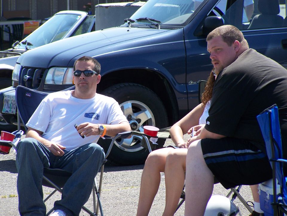 Three people sit in camping chairs outdoors, with a dark blue SUV and a lighter colored convertible parked behind them