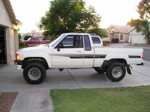 1986 Toyota Hilux - Side view of a white 1986 Toyota Hilux pickup truck parked in front of a garage, with another truck behind it.