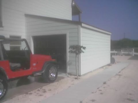 1989 Ford Bronco - A red Jeep Wrangler sits parked beside a white garage on a sunny day.