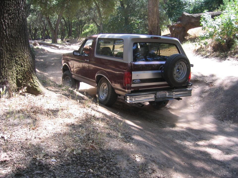 Maroon and tan Ford Bronco driving on a dirt road, kicking up dust. Spare tire mounted on the rear