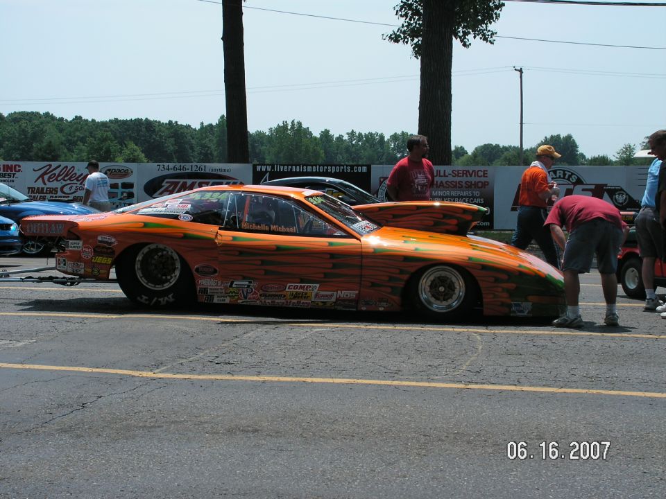 Orange drag car with green stripe graphics and numerous sponsor decals, parked on asphalt with people around