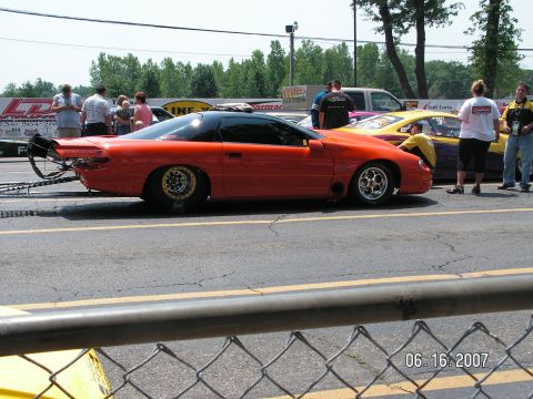 1988 Ford Mustang - An orange drag car with a black roof sits on a race track, with other vehicles and spectators in the background.