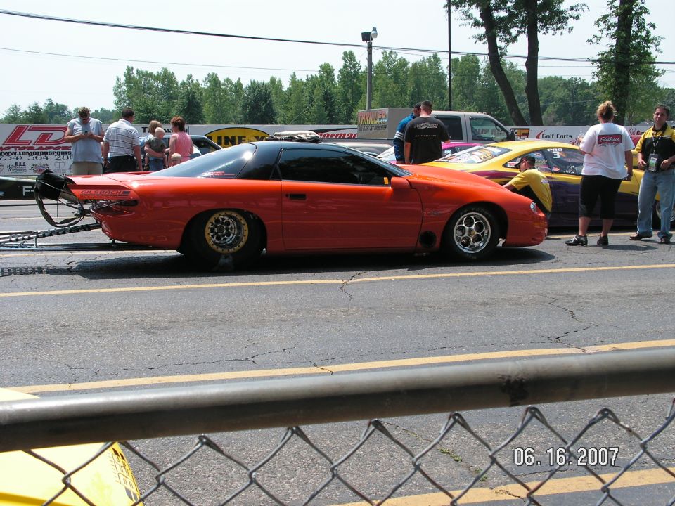 Orange drag car with a black roof and large rear spoiler, parked on a race track