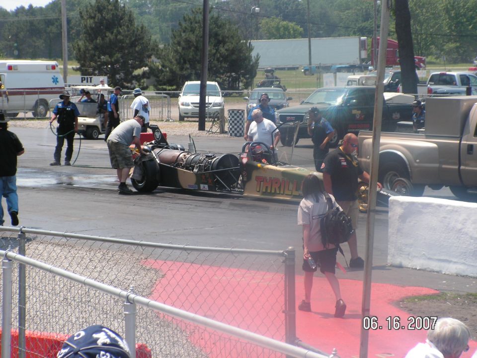 Dragster with jet engine and camouflage paint on track, people around. Pickup truck and ambulance in background