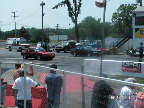 1988 Ford Mustang - A red coupe with a hood scoop is on the drag strip, with an ambulance and other vehicles in the background.