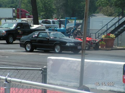 1988 Ford Mustang - A black 1988 Ford Mustang is seen on the race track, with other vehicles in the background.