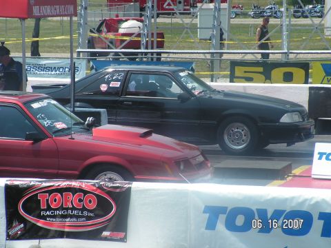1988 Ford Mustang - Two drag cars, a red one and a black one, are lined up on a drag strip.