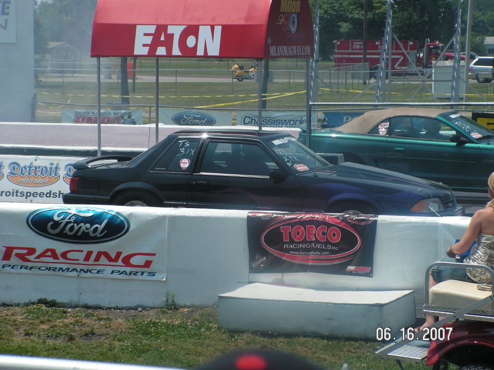 Black Ford Mustang coupe with a spoiler and aftermarket hood on a drag strip