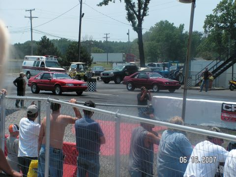 1988 Ford Mustang - A red 1988 Ford Mustang competes in a drag race, with an ambulance and other vehicles visible in the background.