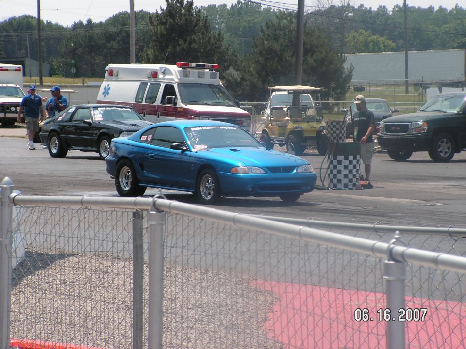 Blue coupe with racing slicks on a drag strip, an ambulance and other vehicles in the background