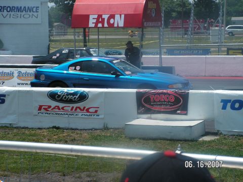 1988 Ford Mustang - A blue Ford Mustang is staged at the drag strip, with another car behind it and racing banners visible.