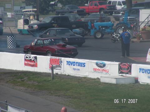 1988 Ford Mustang - A red 1988 Ford Mustang drag car with a prominent hood scoop is lined up on the track, with other vehicles and spectators in the background.