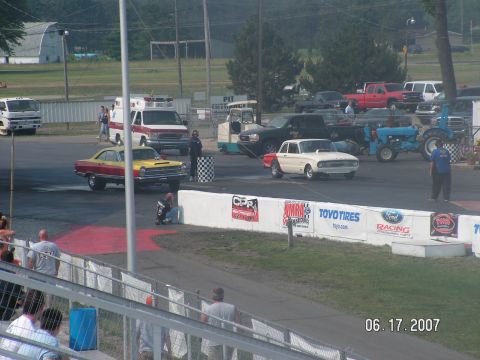 1988 Ford Mustang - A white 1960s Ford Falcon is positioned at the starting line of a drag strip, with other vehicles and spectators visible in the background.