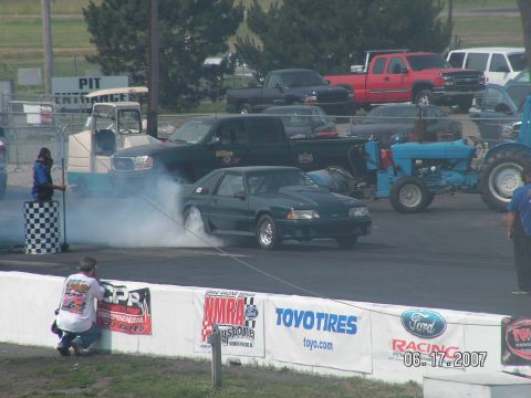 1988 Ford Mustang - A green 1988 Ford Mustang performs a burnout at a drag racing event, with spectators and other vehicles present.