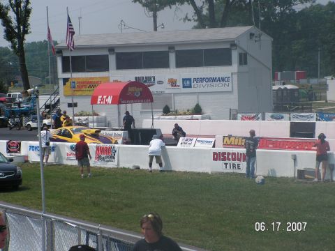 1988 Ford Mustang - A drag racing car is positioned on the track in front of a building with sponsor banners.
