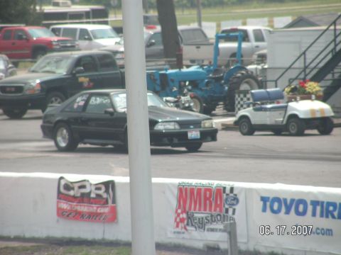 1988 Ford Mustang - A black Ford Mustang is on the drag strip, with other vehicles and a tractor visible in the background.