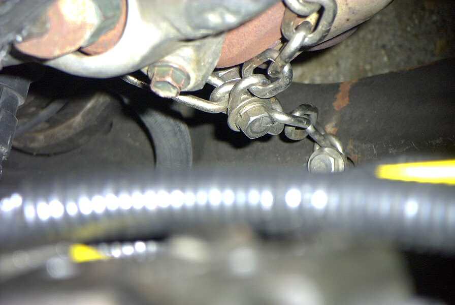Underbody close-up of a plated retention chain bolted between brackets near flex conduit