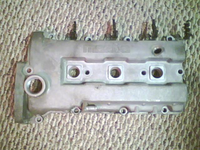 Aluminum engine valve cover with 'MAZDA' embossed on top. Four spark plug holes are visible
