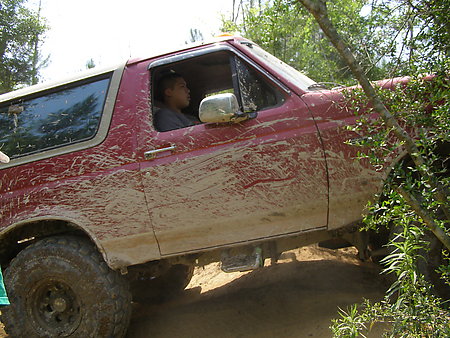 1995 Ford Bronco - Side view of a red two-door Ford SUV caked in dried mud along the doors and rocker panels, tilted at an angle on a rutted dirt trail. The d…