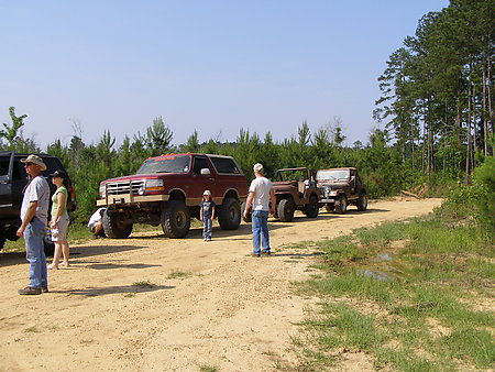 1995 Ford Bronco - A group of off-road vehicles, including a 1995 Ford Bronco and two Jeeps, are parked on a dirt road surrounded by trees.