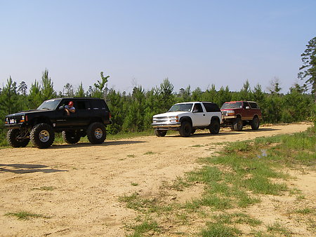 1995 Ford Bronco - Three SUVs are driving on a dirt road with trees in the background.