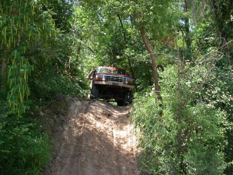 1995 Ford Bronco - A red SUV navigates a steep, muddy trail surrounded by dense trees.